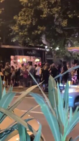 A group of people are gathered outside a building at night. The building appears to be a bar or restaurant, with lights visible inside. Some people are standing in line, while others are interacting with each other. A car is driving by in the foreground, and plants are visible in the bottom of the frame. The scene is illuminated by artificial lights.