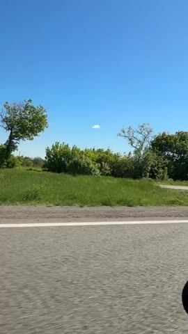 The video captures a side-view from a moving vehicle, showcasing a rural landscape under a clear blue sky. The foreground features a grey asphalt road with a white line, indicating motion. Beyond the road, there's a strip of green grass, followed by a dense line of trees and bushes. The trees are fully leafed, suggesting spring or summer. The sky is bright and clear, with only a tiny, distant white cloud visible briefly around the 0:02 mark. The overall scene depicts a sunny day with typical roadside scenery, likely in the area of Kam'yana Yaruha, Ukraine. There are no people or visible text. A prominent wind noise is audible throughout the recording, suggesting the vehicle is moving at speed or a window is open.