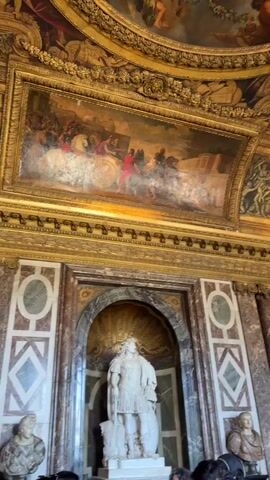 This video shows the interior of a lavishly decorated room, likely a palace or grand hall. The ceiling is adorned with intricate frescoes depicting mythological scenes with cherubic figures and ornate golden trim. Below the ceiling, a large, rectangular painting is mounted, featuring a historical or mythological scene with figures in classical attire, including a chariot pulled by horses. The walls are decorated with marble panels and geometric designs, and feature ornate gold detailing along the ceiling line. Centered in the lower part of the frame, within a recessed alcove, stands a white marble statue of a male figure in Roman-esque armor, holding a shield. The statue is positioned in front of a large, golden shell-like ornament. To the right of the statue, part of another bust or statue is visible. The lighting appears to be bright, illuminating the rich details of the architecture and artwork.