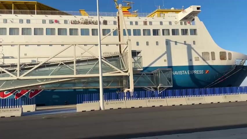 A passenger ferry named Navista Express is docked at a port. A gangway extends from the shore to the ship, allowing passengers to board. The ferry is white with blue accents and has multiple decks with windows. Lifeboats are visible on the side of the ship. The port area has concrete barriers with the text "Puerto de Huelva" visible. The sky is clear and blue, indicating a sunny day. The video is shot from a slightly low angle, looking up at the ferry.