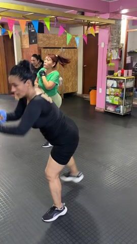 A group of women are participating in a dance fitness class in a studio. The main focus is on a woman in the foreground wearing a black long-sleeved leotard and black shorts. She is actively moving her arms, holding blue hand weights, and appears to be following along with the instructor. In the background, other women are also engaged in the exercise routine, some in athletic wear. The studio environment is decorated with colorful triangular flags hanging from the ceiling. There are wooden cabinets and shelves with various items, including products that appear to be health or beauty related. The floor is a dark, textured mat. The lighting is bright, illuminating the space.
