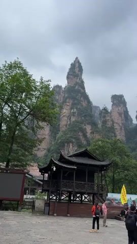 The video shows a crowd of people gathered in a paved square in front of towering rock formations covered in lush green vegetation. The sky is overcast. To the left of the crowd is a traditional Chinese-style wooden pavilion. A yellow flag with Chinese characters is visible near the center of the square, behind a banner with Chinese writing. Most of the people are standing and looking at the rock formations, while a few are sitting on the steps. They are dressed in a variety of casual clothing.  The trees surrounding the square are dense and green, contributing to the overall natural setting.