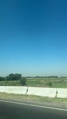 The video shows a view from a moving vehicle, likely a car, looking out over a roadside barrier. The sky is clear and blue. On the other side of the barrier are fields and trees. The barrier itself is made of concrete and appears to be a highway divider. There are some small poles with what look like solar panels or lights visible on the right side of the barrier. The overall scene suggests a rural or semi-rural environment on a sunny day. The audio appears to be music.