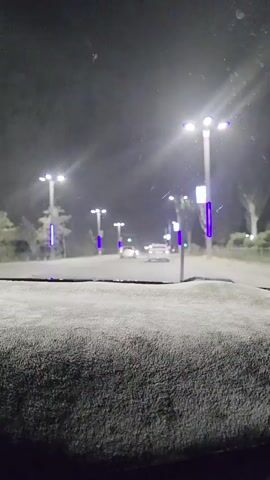 The video shows a view from inside a vehicle driving at night. The road is illuminated by streetlights, some of which have purple accents. There are several cars on the road ahead. The windshield appears to be wet or dusty, with some smudges visible. The overall atmosphere is dark and hazy, possibly due to weather conditions or dust in the air. The camera appears to be mounted on the dashboard or in front of it, as a textured surface is visible in the foreground. There are conversations happening in the background, but they are unintelligible.