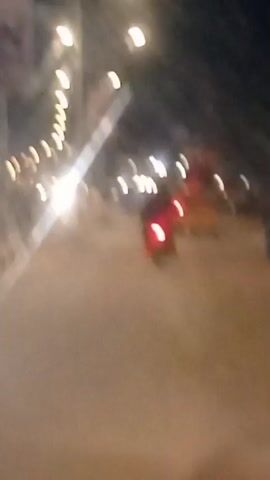 The video displays a night scene on a street. There are streetlights visible on the left side of the frame, casting a glow on the road. In the center of the frame, a red taillight of a motorcycle is prominent, indicating it is moving away from the camera. The motorcycle appears to have a rider, but their features are indistinct due to the motion blur and low light. Further down the road, other vehicles and lights are visible but are out of focus. The overall impression is that of a moving vehicle recording the scene from behind another vehicle at night, possibly in a city or town. The road surface is paved, and there are faint outlines of trees or structures along the side of the road.