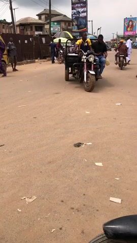 The video shows a dusty street with a few vehicles and pedestrians. In the foreground, a black motorcycle wheel is visible. The camera is positioned low, looking up at the scene. In the midground, a three-wheeled cargo motorcycle is driving away from the camera, carrying two men and some cargo. To the left, a motorcycle with two riders is also moving away. Further down the street, there are several other motorcycles and people walking. The street is lined with buildings and signs. The overall impression is a busy street scene in an urban or semi-urban environment.