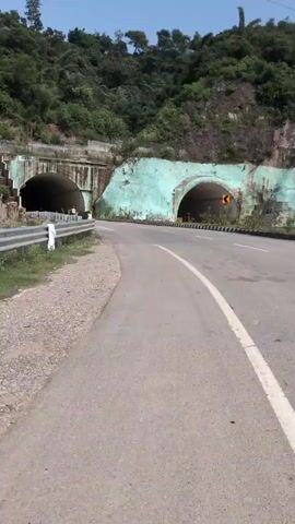 The video shows a two-lane asphalt road curving to the right, leading towards two tunnels carved into a lush green mountainside. The tunnel on the left is dark and appears to be the primary entrance, with a person on a bicycle visible inside. The tunnel on the right is slightly smaller and painted a light blue, with a yellow directional arrow indicating a turn. Guardrails line the left side of the road, and the slopes of the mountain are covered in vegetation. The sky is clear and blue.