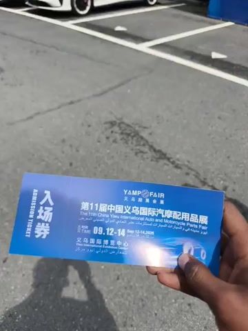 The video shows a person standing in a parking lot holding an admission ticket to the 11th China Yiwu International Auto and Motorcycle Parts Fair. The ticket is blue and white with Chinese and English text. Behind the person is a large blue sign that reads "2026 CHINA YIWU INTERNATIONAL AUTO & MOTORCYCLE PARTS EXPO" in English and Chinese. The sign also indicates the dates of the expo as March 31-April 2, 2026. A tall building can be seen in the background. The person moves towards the sign. A white car is parked in the lot.