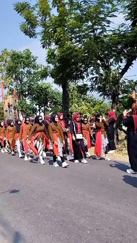 A group of people, primarily women wearing hijabs and traditional clothing, are walking in a procession on a paved road. They are dressed in brown tops, colorful patterned skirts, and red sashes or wraps around their waists. Some are wearing white shoes, and others appear to be wearing sandals. The procession is moving from left to right in the frame. The environment is outdoors, with trees and foliage visible on either side of the road. The sky is blue with some clouds. In the foreground, a person with a light-colored hijab and a burgundy top is walking away from the camera, holding a red object in their hand. The overall scene appears to be a cultural event or parade.