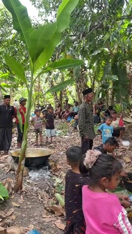 A group of people, a mix of adults and children, are gathered in an outdoor, wooded area. In the foreground, a large pot sits over a fire, appearing to be used for cooking. Several children are in the immediate foreground, some facing away from the camera. Adults are seated and standing in the background, some engaged in conversation. One man, wearing a patterned shirt and a cap, stands facing left. Another man, wearing a black shirt and pants, stands on the left side of the frame. The environment is lush with trees and foliage. The ground is covered in dirt and leaves.