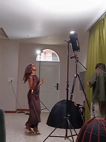 The video shows a woman modeling in a room with photography equipment. The woman is wearing a brown sleeveless dress and gold-colored high heels. She has long brown hair and is wearing sunglasses. She is standing in the middle of the room, posing for the camera. A photographer stands to her right, operating camera equipment. 

The room is sparsely furnished with white walls. There are two photography lights on tripods, one behind the model and another next to the photographer. The photographer's light is rigged with a black umbrella. A green curtain hangs along the right side of the frame. A white door is visible in the background, leading to another room.