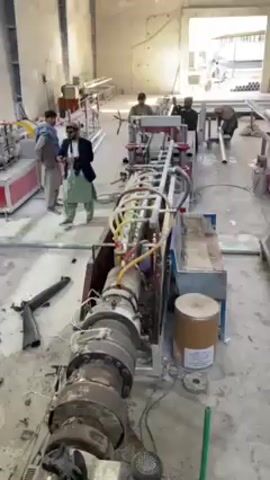 The video shows an industrial setting with several men operating machinery that appears to be involved in pipe production.  

One man in a suit and traditional clothing walks in the foreground, holding a cell phone. Another man appears to be adjusting a machine. There are several machines present; one long machine with multiple connected cylindrical components, connected to an extrusion process on the right, with a barrel of material near it. Other men are handling or inspecting the finished pipes at a cutting machine down the production line. A black rifle or long gun lies on the floor near the front of the primary machine in the foreground. The floor appears to be concrete and is somewhat dirty. The setting is an open building, with a doorway visible in the background.