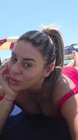 A woman with blonde hair in a ponytail, wearing a pink swimsuit, is lying on a towel by a body of water. She is looking at the camera and resting her chin on her hand. In the background, there are structures that appear to be part of a resort or amusement park, including a Ferris wheel and a roller coaster. The sky is blue and the sun is shining.