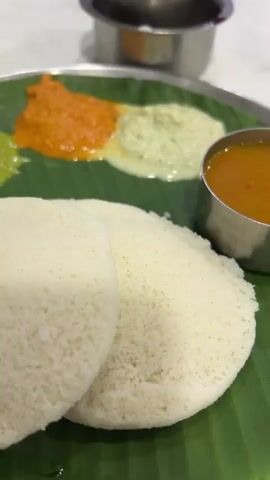 The video shows a close-up view of a plate of idli served on a banana leaf. The plate contains two idlis, three chutneys (green, orange, and white), and a small bowl of sambar. In the background, there are other plates, cups, and people in a restaurant setting. The lighting is bright, and the camera is focused on the food.