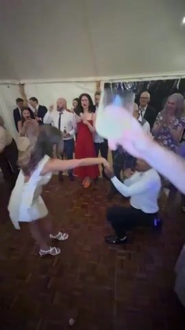 This video shows a proposal happening at a party. A man is on one knee, holding out a ring to a woman who is dancing. They are surrounded by other people who are watching and cheering. The environment appears to be a tent with a wooden dance floor. The lighting is dim, with some colored lights adding to the festive atmosphere. The woman is wearing a white dress and the man is wearing a white shirt and black pants.