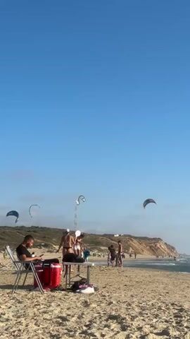 The video shows a beach scene under a clear blue sky. In the foreground, on the sandy beach, are several people. A man sits in a folding chair on the left, looking down at a red cooler and a table. A few other people are standing near the table, and some are walking further down the beach.

In the middle ground, the ocean stretches out, with moderate waves breaking on the shore. Several kitesurfers are visible in the distance, with their kites in the air over the water. The coastline features sandy dunes and some vegetation.

The overall impression is a sunny day at the beach with activities like kitesurfing taking place.