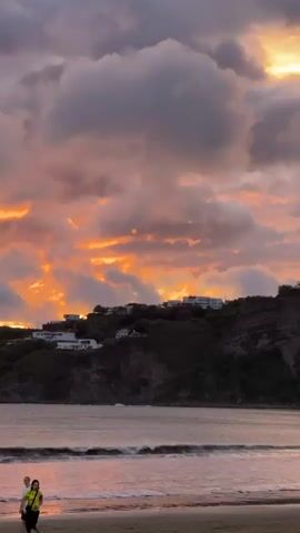 The video shows a tranquil beach scene at sunset. The sky is filled with dramatic, brightly colored clouds, illuminated by the setting sun. Below the sky, a dark, forested hill is dotted with white buildings. The sea is calm, with gentle waves lapping at the shore. In the foreground, on the sandy beach, two people are walking near the water's edge. One person is wearing a yellow shirt and the other a light-colored shirt. The overall mood is peaceful and serene.