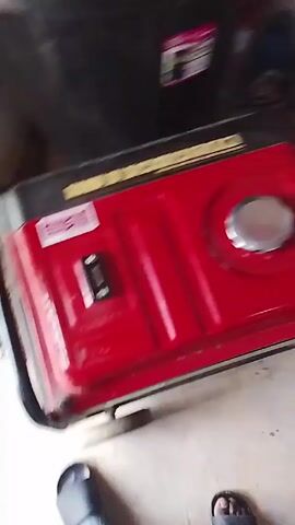 The video shows a red gasoline generator sitting on a concrete floor. The generator has a black frame and a silver gas cap. There is a label on the side of the generator and a switch. To the left of the generator, there is a black plastic container. A pair of sandals are visible near the bottom of the frame. The camera is slightly shaky and moves slightly during the video. The lighting is somewhat dim.