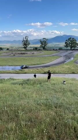 A black sports car drifts around a corner on a paved track, creating a large cloud of white smoke. Two people and a dog are standing in a grassy field in the foreground, watching the car. In the background, there are trees, mountains, and a clear blue sky with scattered clouds. The scene appears to be a sunny day.