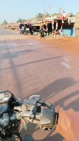 THE VIDEO SHOWS A FIRST-PERSON PERSPECTIVE FROM A MOTORCYCLE RIDER DRIVING DOWN A DIRT ROAD IN A RURAL VILLAGE. THE RIDER'S HANDS ARE VISIBLE ON THE HANDLEBARS. THE ROAD IS LINED WITH SMALL SHOPS AND HOUSES, SOME OF WHICH ARE DECORATED WITH INDONESIAN FLAGS. THERE ARE SEVERAL MOTORCYCLES AND A SMALL TRUCK PARKED ALONG THE ROAD. PEOPLE CAN BE SEEN WORKING AT THE SHOPS. THE SUN IS SHINING, AND THE SKY IS CLEAR.
