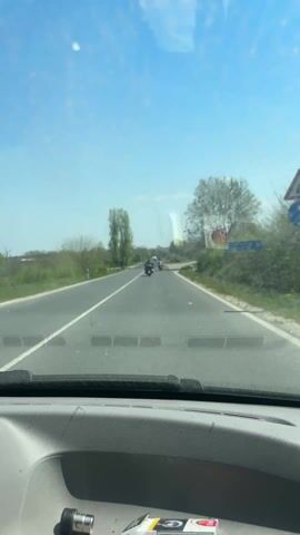 The video shows a view from inside a car, looking out the windshield onto a straight, paved road. There is a white line separating the lanes. Several motorcycles and a white car are traveling in the same direction, away from the camera. The sky is blue with some clouds. Trees and bushes line the sides of the road. A road sign is visible on the right side.