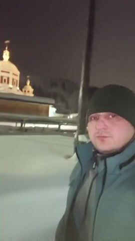 A man in a green beanie and a blue-green jacket is recording a selfie video outside at night. The ground is covered in snow. Behind him is a white and red building with a dome and a cross on top, illuminated by lights. There are also a few flags visible behind the building. The background is dark, suggesting it is nighttime. The man is looking slightly off-center from the camera and appears to be stationary.
