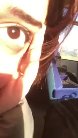A close-up shot shows a person's face from the side, with a brown eye and eyebrow visible. The person is holding a small gold hoop earring near their ear. Some of the person's reddish-brown hair is visible around the face. The background shows a blurry scene, possibly inside a moving vehicle like a bus or train, with a purple seat and other indistinguishable objects. The scene is brightly lit.