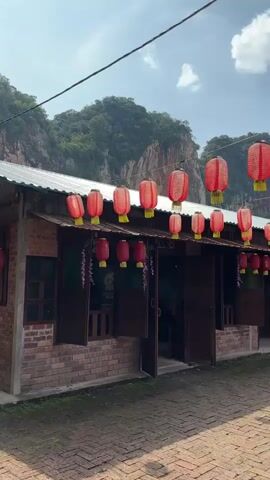 The video pans across a row of traditional brick buildings, decorated with numerous red lanterns strung across wires overhead and hanging from the eaves. The buildings appear to be shops or restaurants, with dark wooden doors and shutters. In the background, a large, rocky mountainside rises up, its slopes covered in greenery. The sky is a bright blue with scattered white clouds. The ground in front of the buildings is paved with red bricks. The overall impression is one of a scenic and culturally rich location, possibly a tourist attraction.
