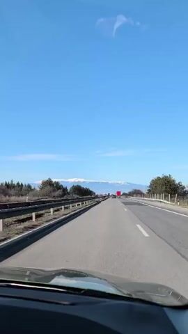 The camera is positioned from inside a vehicle traveling on a multi-lane highway on a sunny day. Cars, trucks, and other vehicles are visible driving in the same direction. A mountain range with snow-covered peaks is visible in the distance. Trees and vegetation line the side of the highway. The sky is clear and blue with minimal clouds.