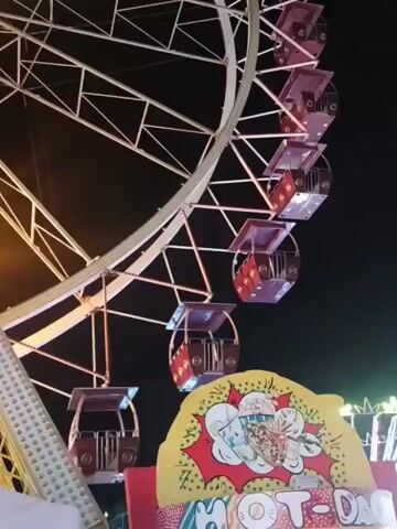 The video shows a Ferris wheel at night. The Ferris wheel has several gondolas, and it is lit up with lights. In the foreground, there is a sign that says "HOT DOGS". The Ferris wheel appears to be in motion.