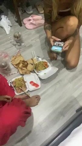 The video shows two women sitting on a light grey floor covered with styrofoam food containers. The woman closest to the camera is wearing a light pink tank top and red pajama pants. She is seated cross-legged and smiling at the camera. Her left arm is tattooed. The other woman is seated on her knees, facing away from the camera, and is holding a cell phone in her hands. The floor is cluttered with various food containers, plastic bags, and a glass. A pair of pink flip flops are visible in the background. A small table with a dark surface and folding legs is also visible in the background.