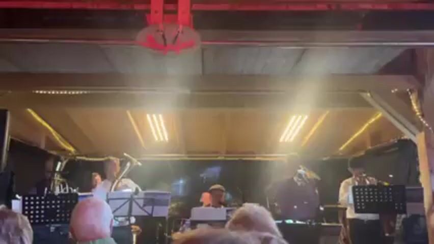 A band of six musicians plays on a stage under a wooden structure. The band consists of a tuba player, a man with a keyboard, and four trumpet players. The players are dressed in smart casual clothing. The venue is enclosed, possibly a bar or small concert hall, with warm lighting and an audience in the foreground. A decorative sign hangs above the stage. The band appears to be performing live music for the audience.