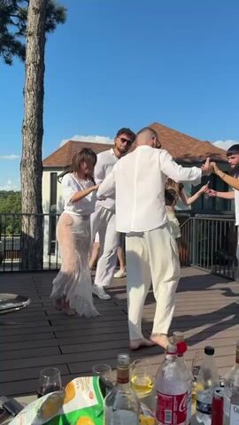 A group of five young adults are dancing in a circle on a wooden deck on a sunny day. The sky is a vibrant blue with a few wispy clouds. The people are all dressed in white or light-colored clothing, suggesting a festive or formal occasion like a wedding or a summer party.

In the foreground, several bottles of alcohol, including Captain Morgan, and some snacks are visible on a table, indicating a relaxed and celebratory atmosphere. The deck appears to be outdoors, possibly on a rooftop or overlooking a body of water, given the glimpse of a railing and greenery in the background. A large tree stands to the left of the frame, adding a natural element to the scene.

The people are engaged in dancing, with smiles and happy expressions on their faces, suggesting joy and camaraderie. The woman on the left is wearing a sheer white lace dress over white undergarments, which adds a touch of elegance and perhaps a bohemian flair to her attire. The overall mood is lighthearted and joyful. The scene takes place during the daytime, likely late afternoon given the warm lighting and long shadows. The location context provided is Cojușna, Moldova.