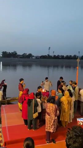 A group of people are gathered on a red platform next to a body of water. The sky is overcast and appears to be dusk. There are streetlights visible in the background across the water, and what looks like a stadium with lights in the distance. In the foreground, a few people are holding up cell phones to record the scene. At the center of the gathering, a fire is burning on a small altar-like structure. Several individuals are standing around the fire, with some appearing to be participating in a ritual or ceremony. People are dressed in casual clothing, with some individuals wearing traditional Indian attire such as kurtas and sarees. The overall atmosphere seems to be one of communal gathering and observance.