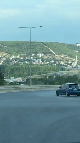A blue Volkswagen Golf is driving on a highway. The car is in the right lane, and the camera is filming from another vehicle in the left lane. The highway is elevated, with a grassy slope on the left. In the background, there is a town built on a hillside, with buildings of various sizes. A minaret is visible, indicating a mosque. The sky is cloudy, with some patches of blue. The overall impression is one of a suburban or semi-rural landscape.