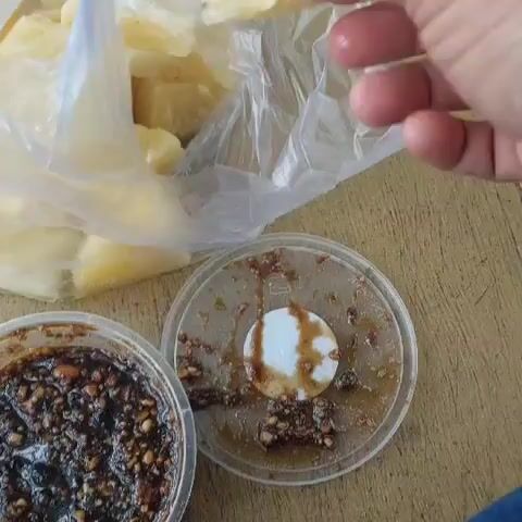 The video shows a close-up view of a person's hand dipping a piece of pineapple into a small container of dark brown sauce. The sauce appears to have small pieces of nuts or seeds in it. In the background, a plastic bag filled with more pieces of pineapple is visible. The scene takes place on a wooden surface, likely a table. The lighting suggests an outdoor or well-lit indoor environment.