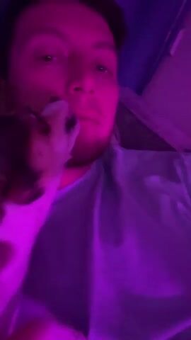 A person with short brown hair and a beard is illuminated by pink and purple light. The person is wearing a light blue t-shirt. A small dog, with white, brown, and black markings, is licking the person's face near their mouth. The dog has its paw on the person's cheek. The background appears to be a curtain or wall with vertical lines.