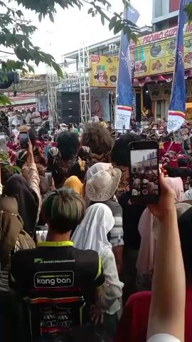 The video shows a crowded outdoor event, likely a street festival or performance. In the foreground, the backs of several people are visible, some holding up phones to record. One person in the center is wearing a black jacket with green lettering. The crowd is dense, with many people wearing headscarves and casual clothing. In the middle ground, a stage or performance area is set up with speakers and a banner that displays images of food. Several performers are visible on the stage, some wearing elaborate, furry costumes that obscure their faces, possibly representing mythical creatures or characters. They appear to be interacting with the crowd or engaged in a performance. In the background, there are buildings with storefronts, including one with a prominent "SONIC CHICKEN" sign, and banners associated with "Bank Jaten" and "PROMO SUPER." The overall atmosphere seems lively and festive, with people gathered to watch the performance. The lighting suggests it is daytime.