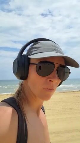 A woman is walking on a beach. She is wearing headphones, a baseball cap, and sunglasses. The sky is blue with some clouds, and the ocean is visible in the background. The sand is light brown.