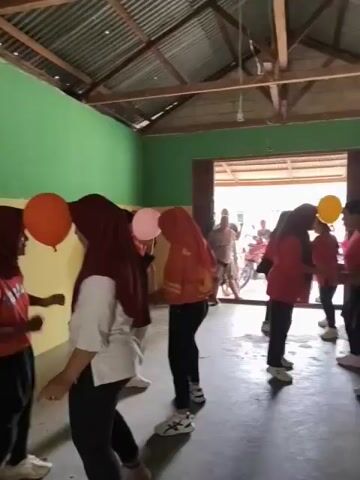 A group of women are playing a game with balloons. The women are wearing hijabs and casual clothing. Some of the women are wearing red shirts and black pants, while others are wearing red shirts with yellow accents. One woman is wearing a white shirt and black pants. The women are standing in pairs and holding balloons between their foreheads. They appear to be trying to keep the balloons from falling. The game is taking place in a room with green and yellow walls and a corrugated metal roof. There are open doorways in the back of the room, and people and a motorcycle can be seen outside.