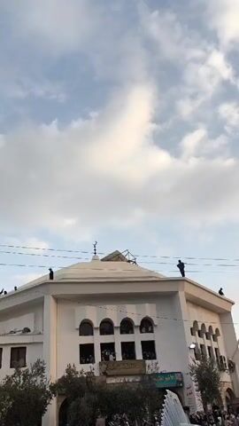 Here's a description of the video:

The video shows a large crowd of people gathered in front of a building. The building appears to be a community center or mosque, adorned with a crescent moon symbol. Some individuals are positioned on the building's roof. The sky above is partly cloudy.

The camera pans to show more of the crowd, revealing people holding Syrian flags and chanting. One man is seen taking pictures with his phone. Uniformed individuals, possibly security personnel, are present within the crowd. Tall, bare trees are visible in the background.  The atmosphere appears to be festive and celebratory.