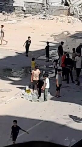 The video shows a group of people, predominantly children and some adults, standing in an outdoor area that appears to be a street or alleyway. There are several large, clear plastic containers on the ground, some appearing to be filled with water. Some children are gathered around these containers. The environment is somewhat barren with a light-colored, possibly dusty, ground and a stone or brick wall in the background. The people are dressed in casual clothing. There are no overt signs of distress or organized activity, other than the collection of water containers. The interactions between the people seem casual, with some standing in groups and others appearing to be observing or waiting. The overall impression is one of everyday life in a potentially resource-scarce environment.