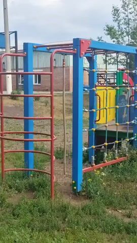 The video shows a playground with various equipment. In the foreground, there's a climbing structure with blue and red poles and a yellow net. To the left, a red ladder is visible. To the right, a portion of a red and green slide is seen. The ground is covered with patches of grass and dirt. In the background, there's an outdoor gym area with several weight machines. A person, wearing a light-colored cap and dark clothing, is seen bending over a piece of gym equipment. Further in the background, there are fences and trees. The sky is partly cloudy. The camera pans across the playground and gym area.