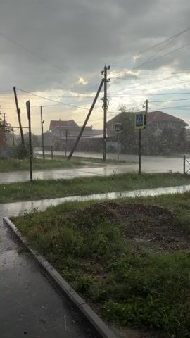 The video shows a street with houses and utility poles during a rainstorm. The ground and road are wet, with puddles forming. Raindrops are visible falling through the air. There are no people or animals visible in the footage.