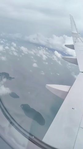 The video shows an aerial view from an airplane window. The right side of the frame is dominated by the wing of the airplane, which is white with some yellow markings. Below the wing, islands can be seen in the ocean, along with scattered clouds. The water appears murky and the sky is overcast. The camera pans slightly to the right, revealing more of the clouds and the vast expanse of the ocean.