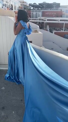 A woman in a flowing blue dress stands on a ledge, her back to the camera. Her dress has a long train that trails behind her. In the background, there is a white building with red accents and several levels of seating or balconies. The sky is clear and blue. The woman raises her hand to her face.