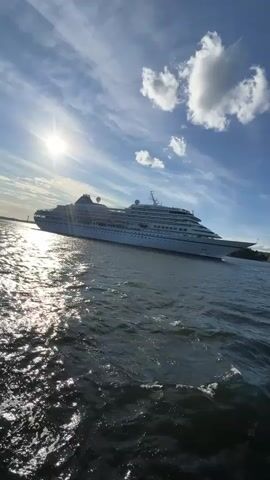 A wide-angle, low-angle, exterior, daytime shot shows a large white cruise ship sailing on the right side of the frame. The sun is visible in the upper left corner, peeking through thin clouds. The water in the foreground is choppy and reflects the sunlight. In the distance, the shoreline is visible with trees and buildings. The sky is blue with scattered white clouds.