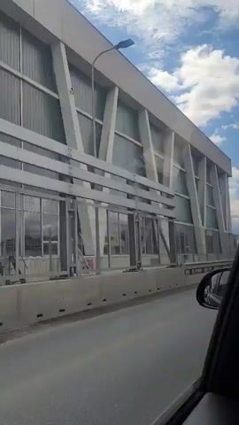The video shows a view from inside a moving vehicle, looking out the passenger side window. The road is visible in the foreground, with a concrete barrier separating it from a large, modern building on the left. The building has a white, angular facade with large glass panels. There are trees and a blue sky with white clouds in the background. A black car mirror is visible in the lower right corner of the frame. The text "Не жди нашего звонка. КПП ЛВ." appears briefly at the end of the video.