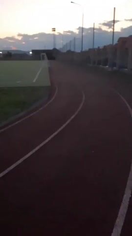 The video shows a view from a running track at dusk. The track is dark red with white lines. To the left of the track is a green field, possibly a sports field. In the background, there are lights, a fence, and buildings. The sky is cloudy and twilight. There are no people or animals visible in the video. The camera appears to be moving slowly, as if being held by someone walking or running.