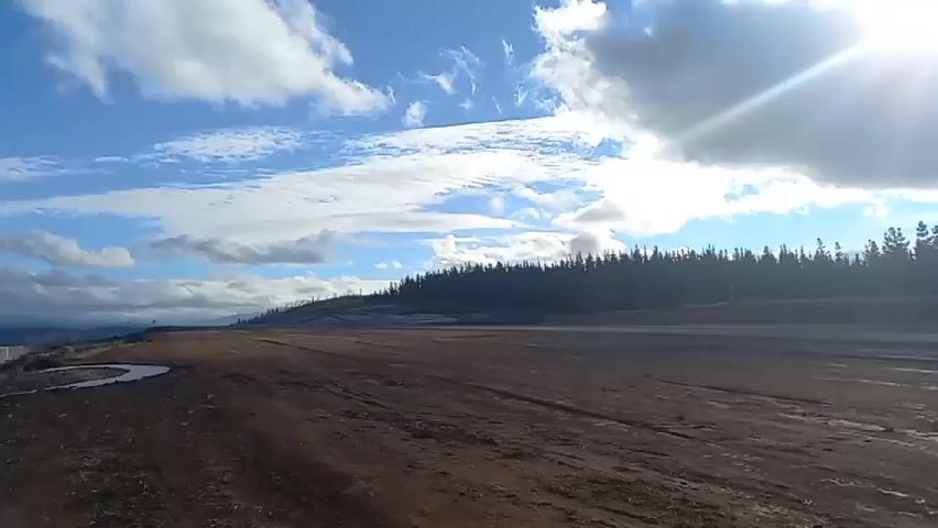 The video shows a landscape of a clear, sunny, slightly cloudy sky above a dirt field that stretches to a dark green line of trees along the horizon. A small puddle is visible on the left. The field has tire tracks and uneven ground. The sun shines brightly from the right.  The camera is stationary.